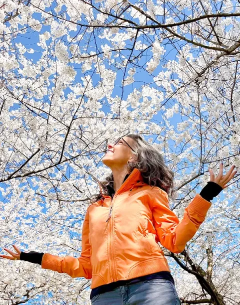Dena in an orange jacket looking up towards a blossoming tree, arms slightly outstretched.