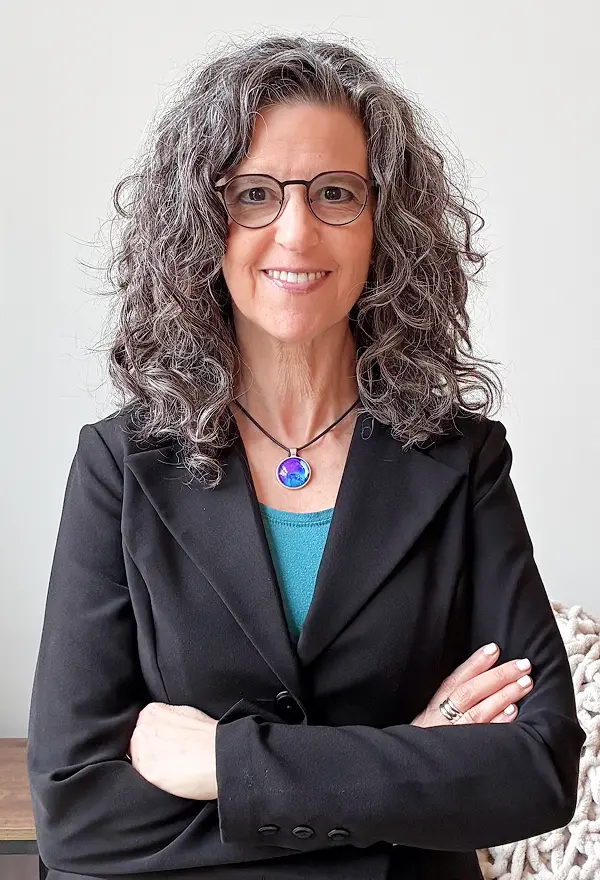 Profile of Dena, in a dark gray blazer with a teal undershirt, arms crossed, with a smile to light up the room.