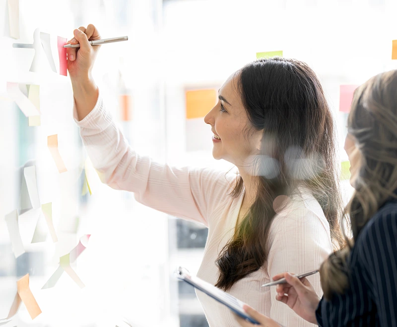Two women write on sticky notes to discuss business objectives